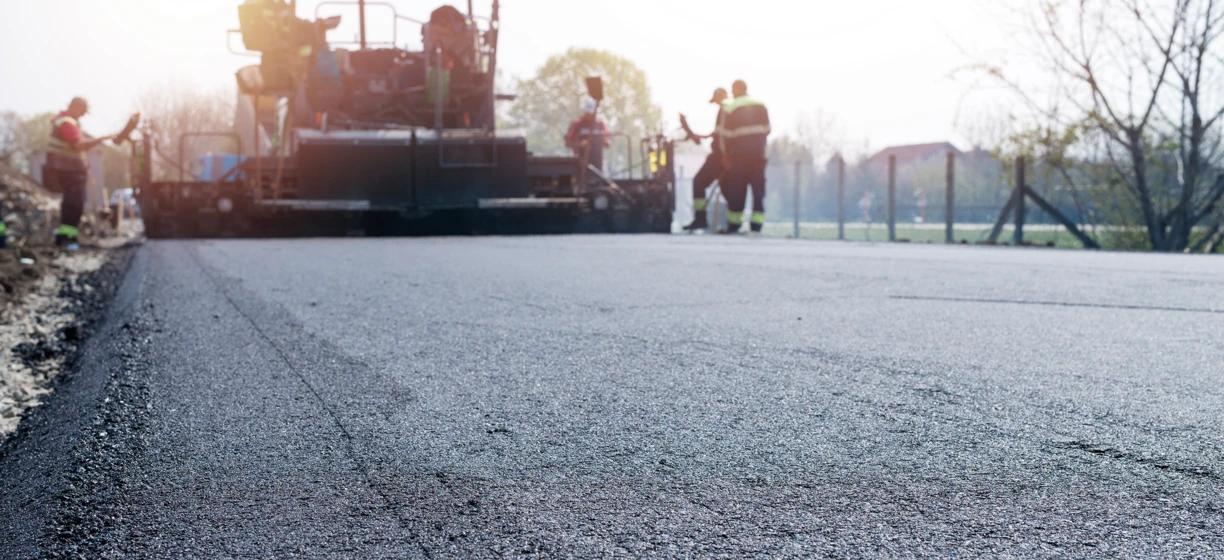 Pracownik fizyczny w odzieży o wysokiej widoczności kładzie asfalt na autostradzie, używając ciężkiego sprzętu. Materiał budowlany wykonany z gumy syntetycznej.