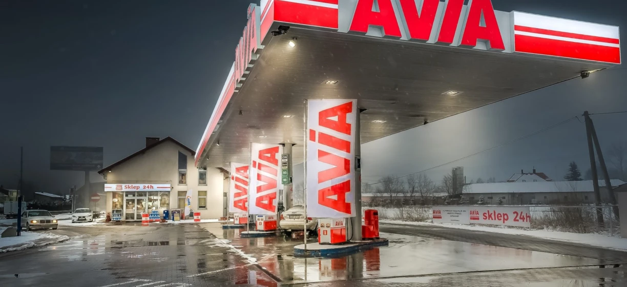 A fuel truck parked at a 24-hour filling station (Sklep 24h) with the brand AVÍA prominently displayed on the bumper.