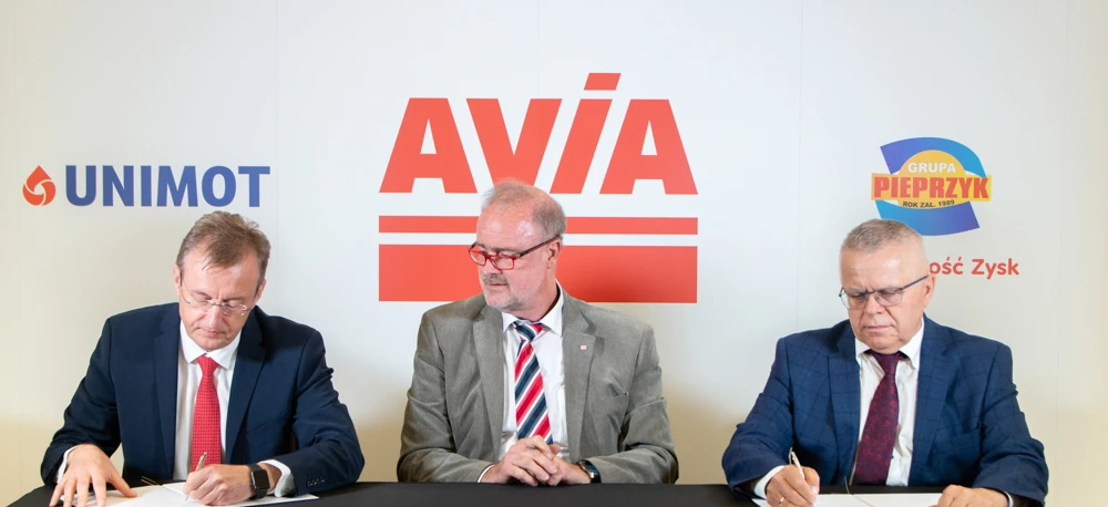Tekst alternatywny: Profesjonalny pracownik umysłowy w garniturze i krawacie, stojący przed reklamowym panelem firmy AVIA. Logo firmy i informacje w języku polskim.