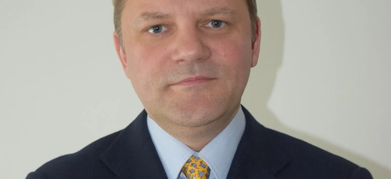 A close-up image of a man in a black blazer and necktie, showing his chin, eyebrow, forehead, and collar as he confidently poses.